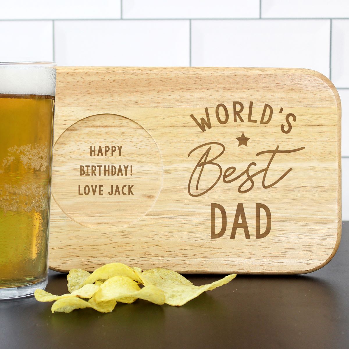 Wooden cutting board with 'World's Best Dad' engraving next to a glass of beer and chips.