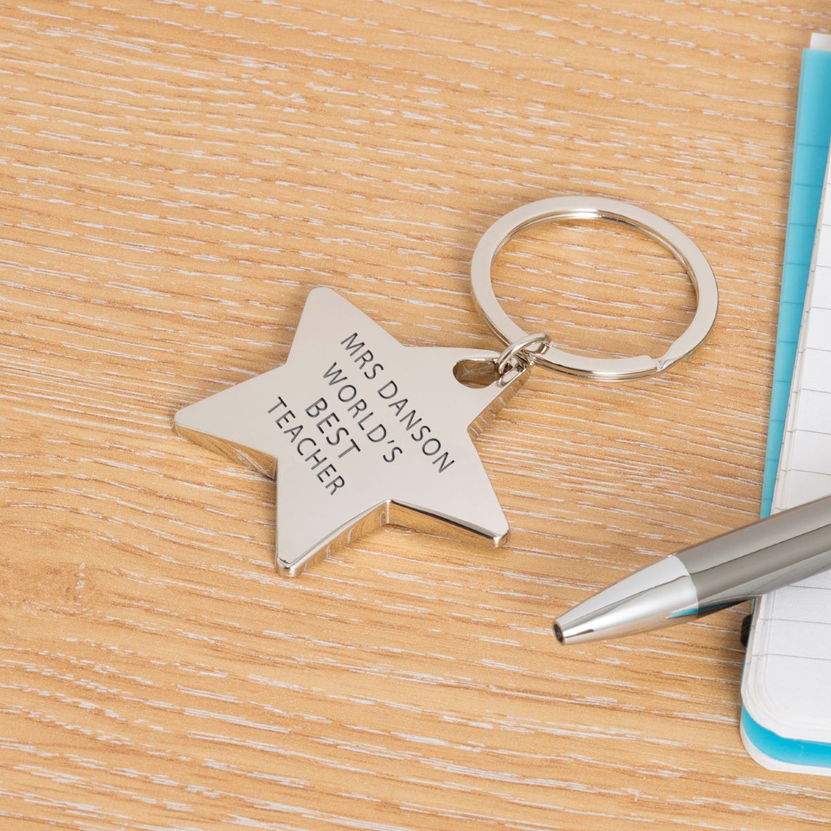 Star-shaped silver keyring with engraved text 'Mrs Danson World's best teacher' displayed on a wooden surface with a pen and notebook.