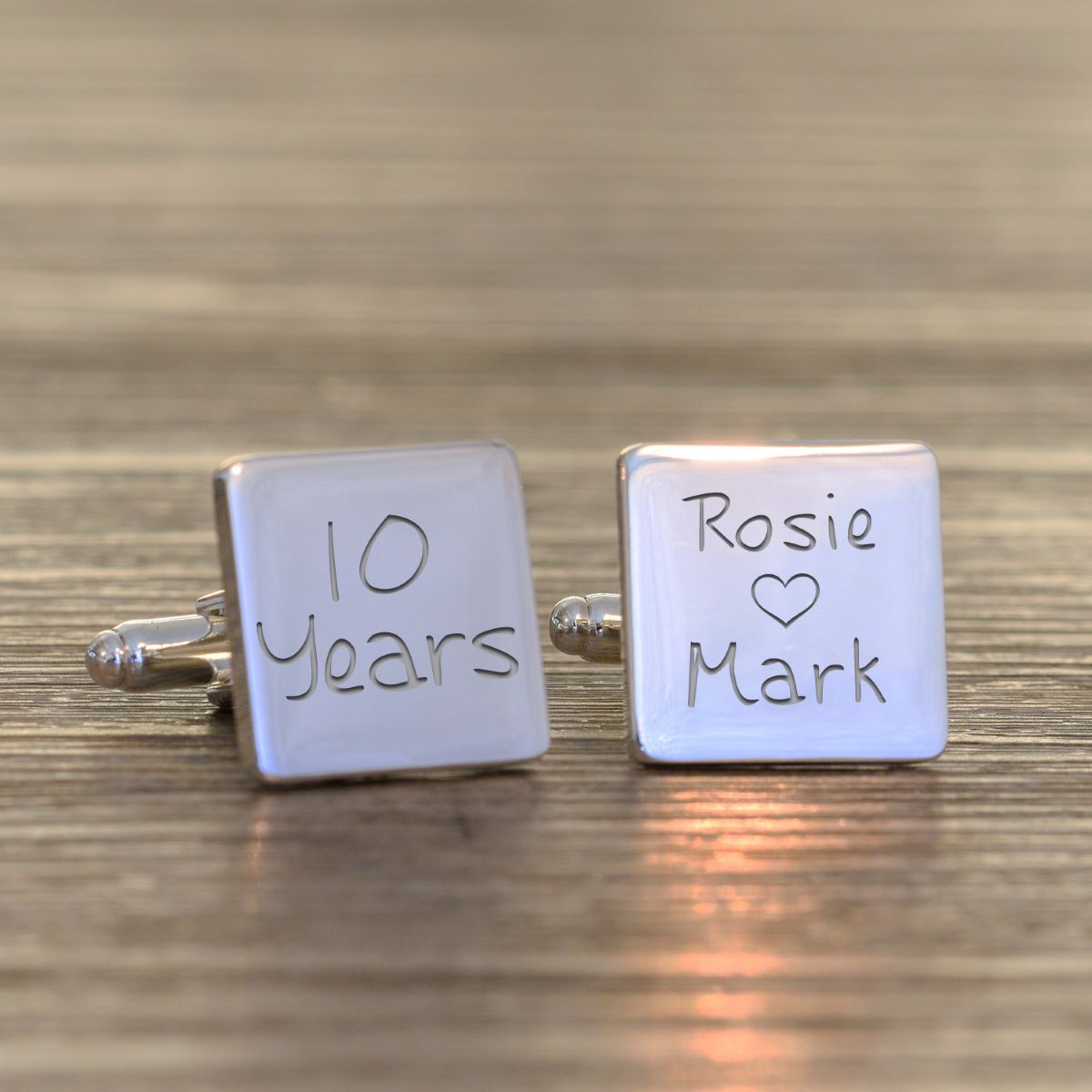 A pair of silver square silver cufflinks engraved with '10 Years' and 'Rosie Heart Mark', displayed on a wooden surface.