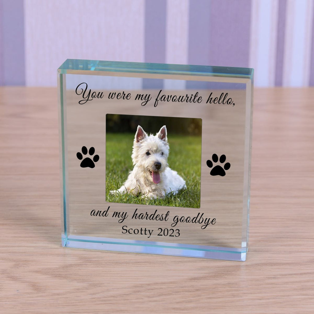 A personalised clear glass block plaque display on a light wood surface, featuring a photo of a white dog, black paw prints, and engraved text reading 'You were my favourite hello. and my hardest goodbye Scotty 2023'.