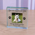 A personalised clear glass block plaque display on a light wood surface, featuring a photo of a white dog, black paw prints, and engraved text reading 'You were my favourite hello. and my hardest goodbye Scotty 2023'.