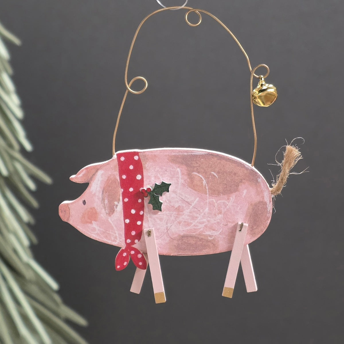 A pink wooden pig ornament hanging from a gold wire loop with a small gold bell and twine tail, decorated with a red polka-dot bow, a small holly leaf, and wooden legs, shown against a dark grey background with blurred greenery at the side.