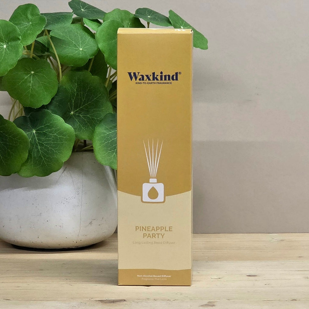 A Waxkind Pineapple Party reed diffuser box in a warm tan and beige color, standing on a light wooden surface next to a potted green plant.