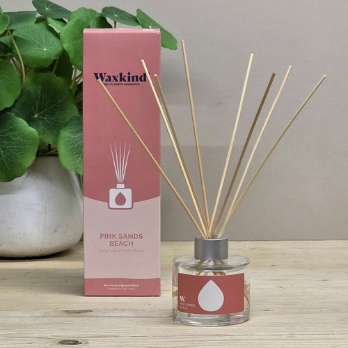 Waxkind Pink Sands Beach reed diffuser with a pink and white box, a glass bottle with a red label, and natural reed sticks, displayed on a wooden table next to a potted green plant.