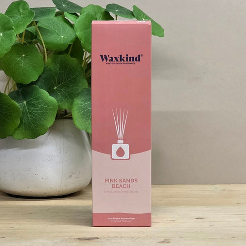 A pink and peach Waxkind reed diffuser box labeled 'Pink Sands Beach' standing on a light wood surface, with a potted green plant in the background.