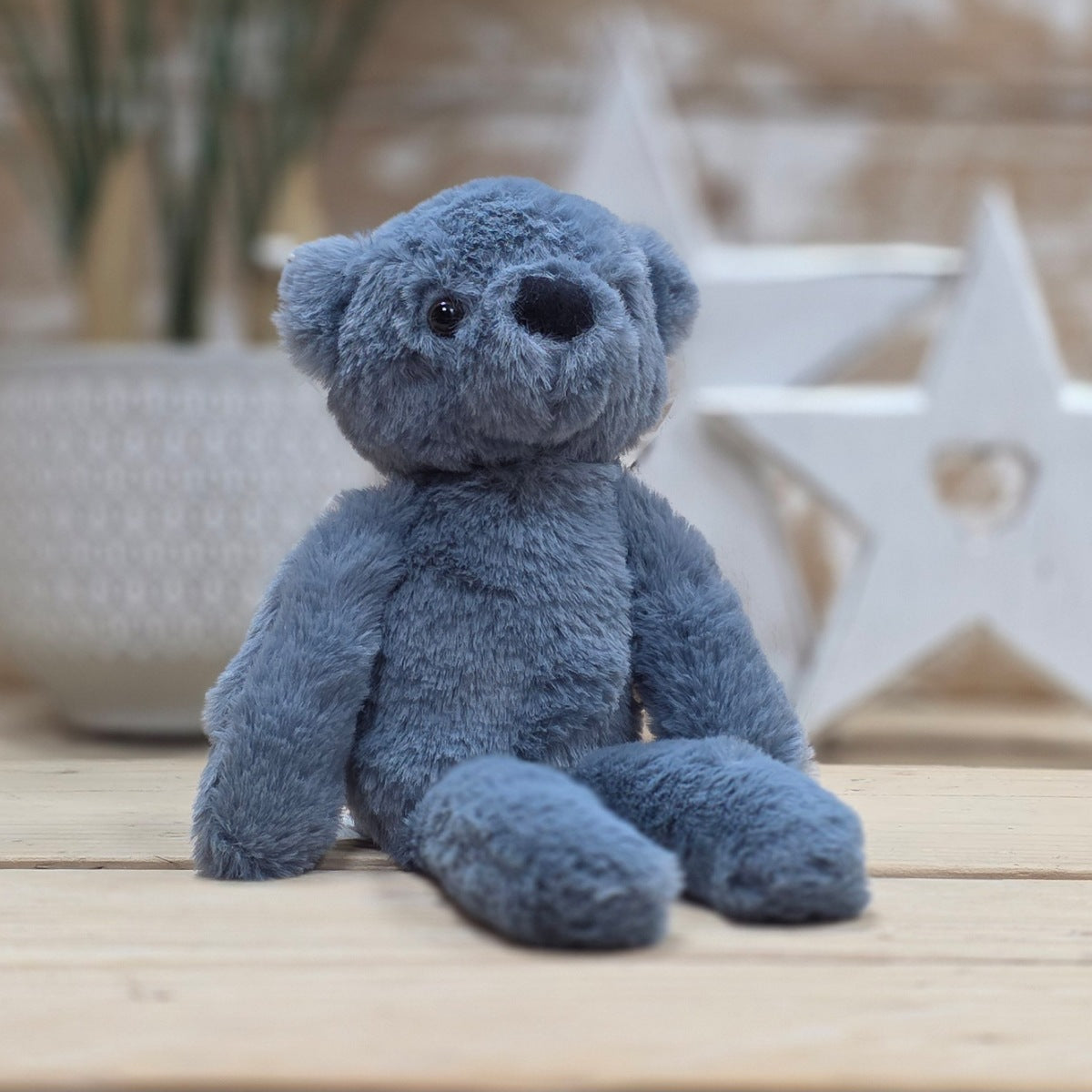 A blue soft fuzzy plush teddy bear toy, with black eyes and nose, on a wooden surface with a blurred star-shaped object in the background
