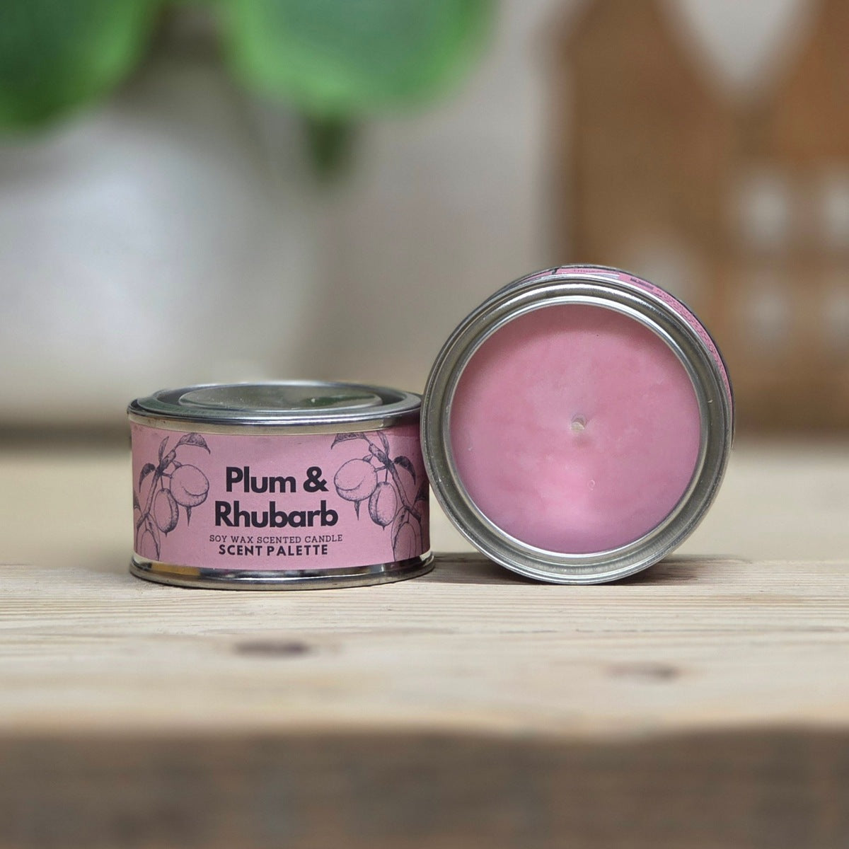 A pink scented candle in a round metal tin with the lid off, placed on a wooden surface. The label reads 'Plum & Rhubarb'.