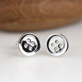 A pair of silver stud earrings shaped like round buttons, resting on a white surface with a blurred brown wooden background.