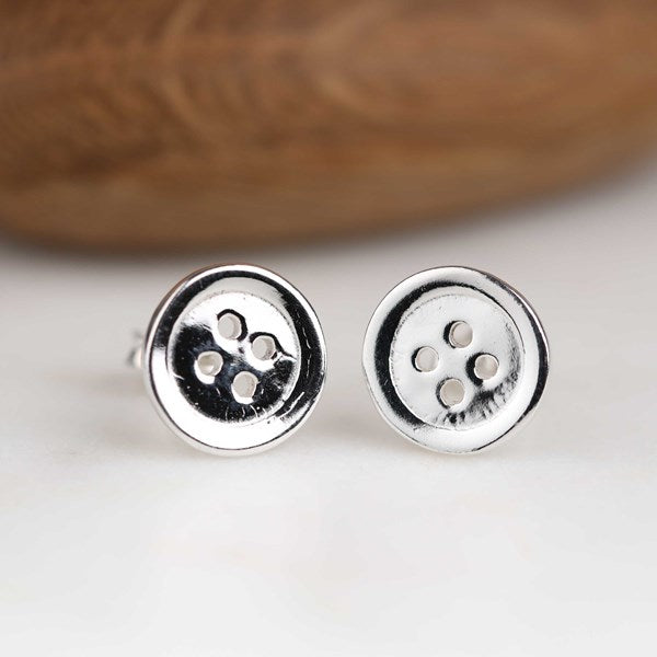 A pair of silver stud earrings shaped like round buttons, resting on a white surface with a blurred brown wooden background.