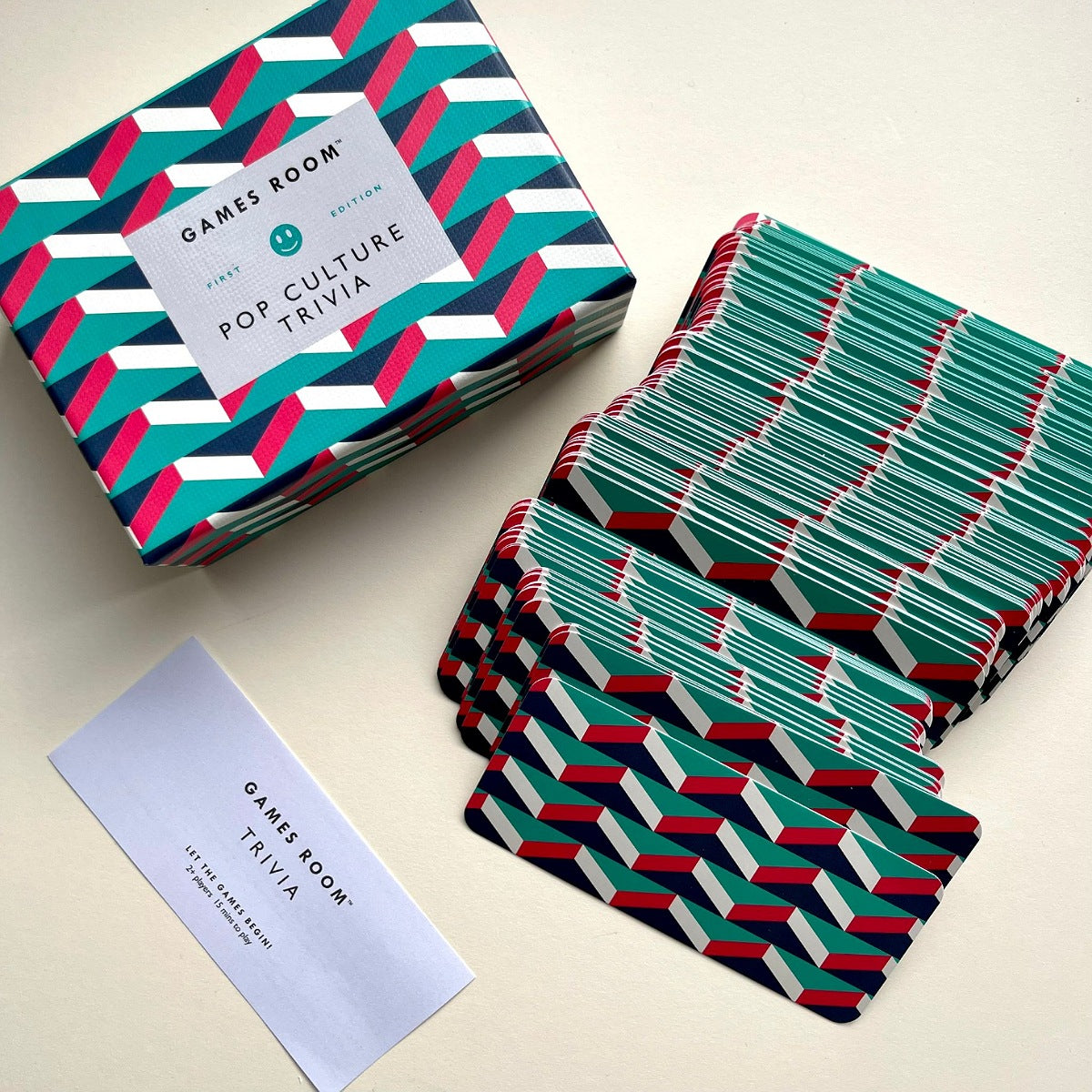 Games Room Pop Culture Trivia deck and box with matching game cards, arranged on a light beige surface. The box and cards feature a bold geometric chevron pattern in teal, pink, white, and navy.