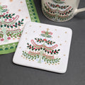 A square white ceramic coaster featuring a festive Christmas tree design with green holly, red berries, and gold stars, displayed on a dark grey surface next to matching napkin and mug.