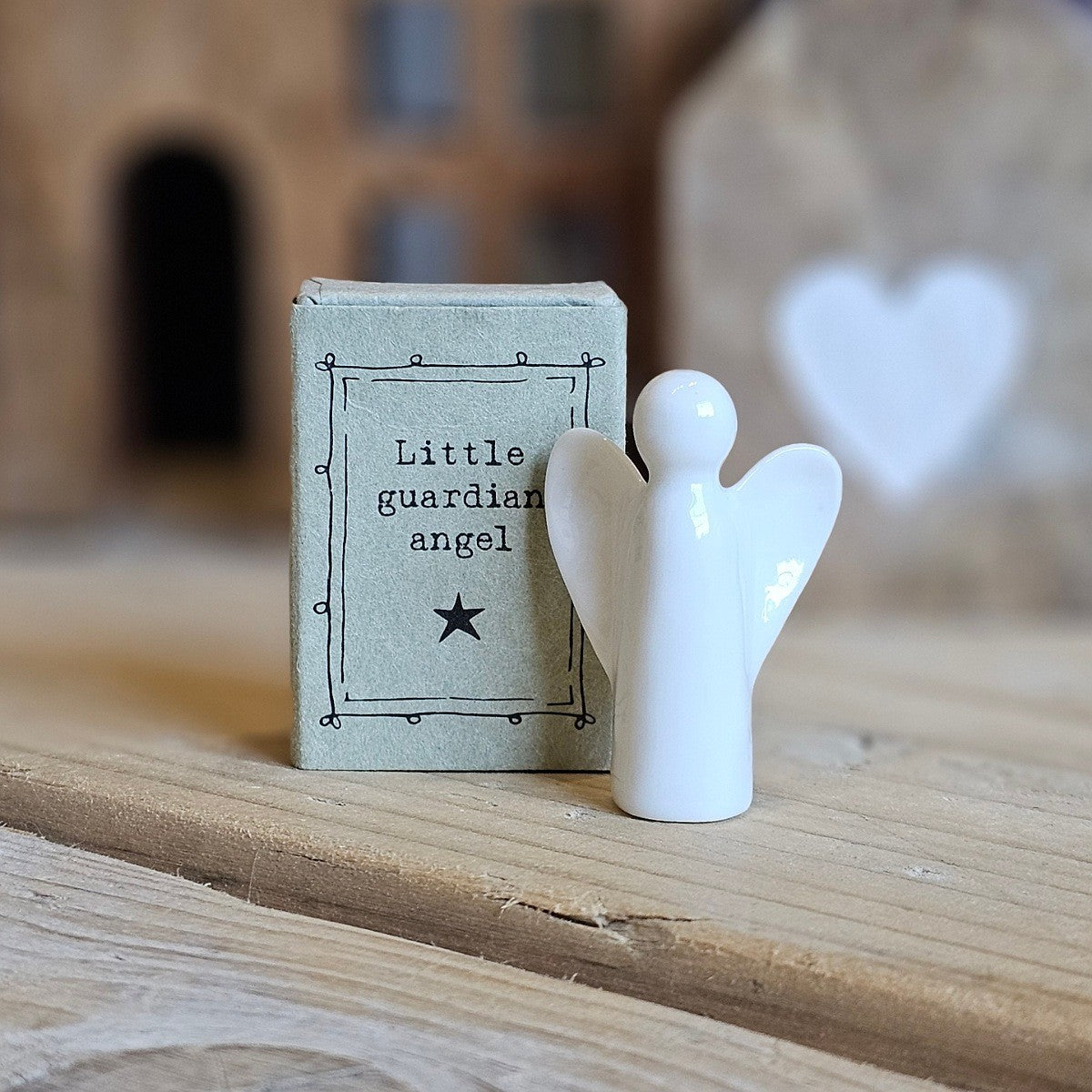 A small white ceramic angel figurine stands beside its light grey packaging box on a wooden surface. The box is printed with the words 'Little guardian angel' and a black star.
