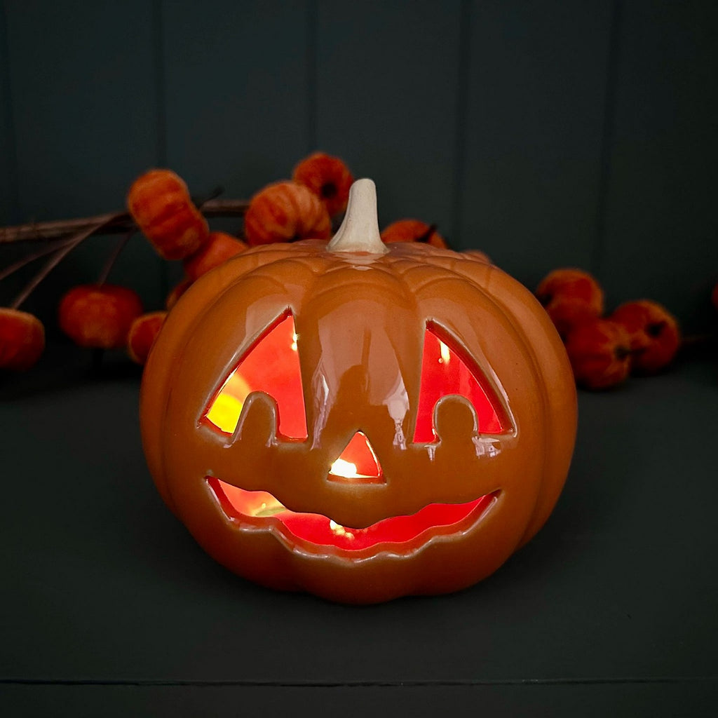 A glowing ceramic orange jack-o'-lantern pumpkin with carved facial features, lit from within with warm red and yellow light, set against a dark background with decorative pumpkins behind it.