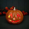 A glowing ceramic orange jack-o'-lantern pumpkin with carved facial features, lit from within with warm red and yellow light, set against a dark background with decorative pumpkins behind it.
