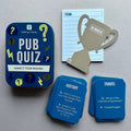 'Pub Quiz' game set on a neutral grey background, featuring a dark blue tin, blue cards, a light blue team notepad, and a gold trophy-shaped winner marker.