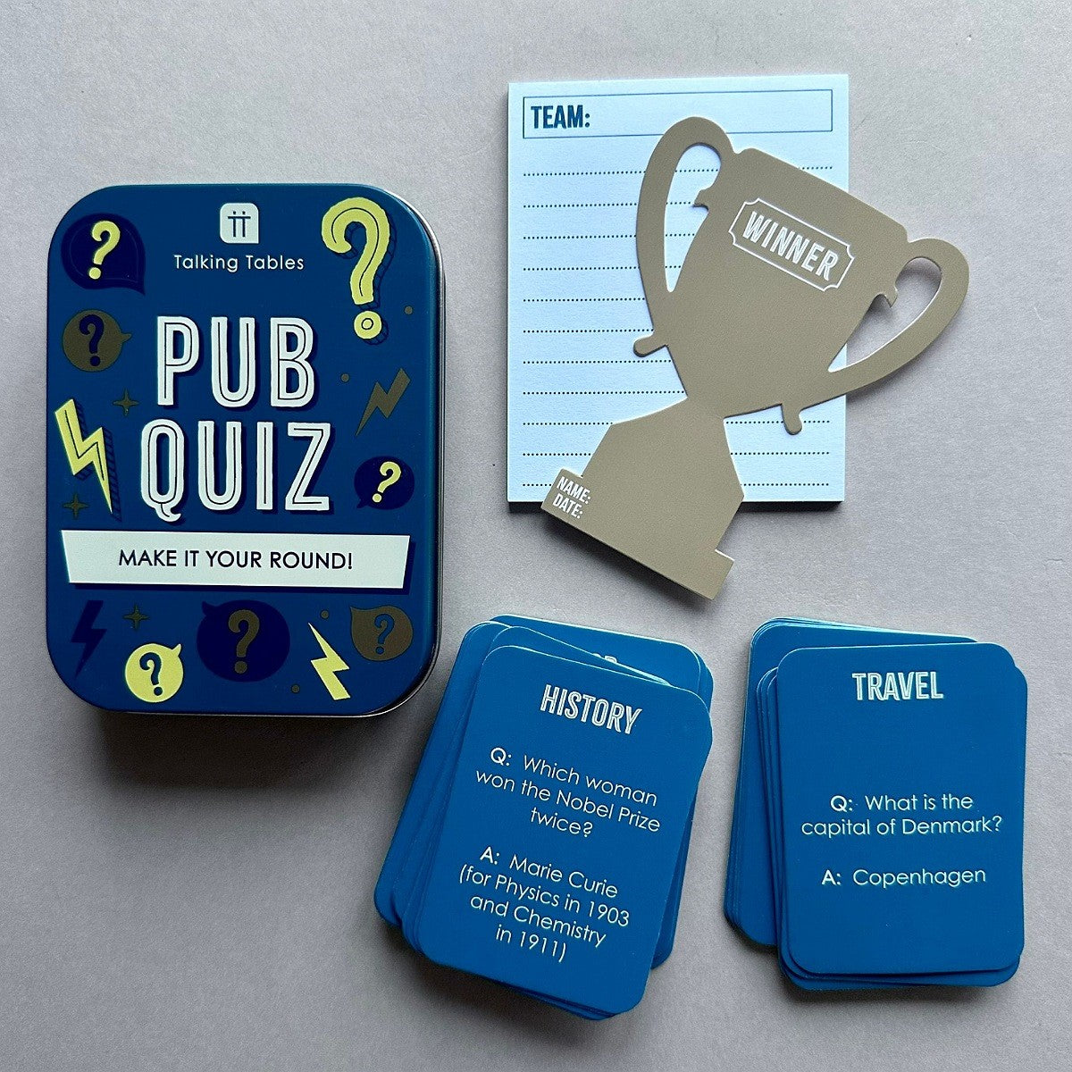 'Pub Quiz' game set on a neutral grey background, featuring a dark blue tin, blue cards, a light blue team notepad, and a gold trophy-shaped winner marker.