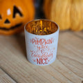 White glass candle holder, gold metallic inside with 'Pumpkin spice & everything nice' text in orange on the front.  With blurred out pumpkins in the background on a wooden surface