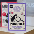 The book cover of 'Purrdle' by Dr Gareth Moore and Laura Jayne Ayres, featuring a purple border, a black cat silhouette, a magnifying glass, and a cream background with puzzle illustrations.