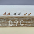 Six small bee-shaped stud earrings in rose gold, silver, and yellow gold finishes, arranged in pairs on a rustic white and wood surface.