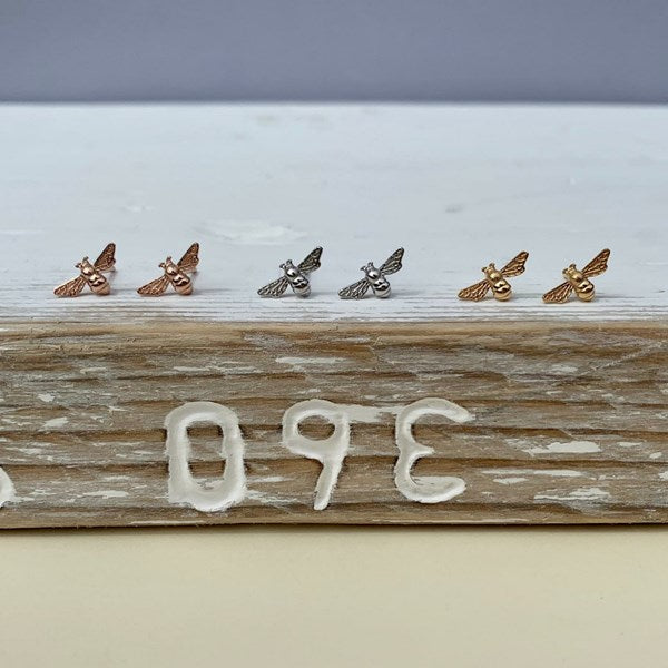 Six small bee-shaped stud earrings in rose gold, silver, and yellow gold finishes, arranged in pairs on a rustic white and wood surface.