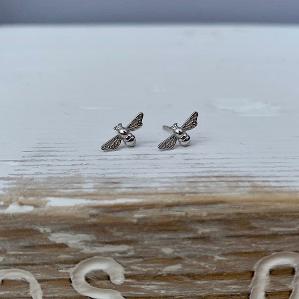 A pair of small silver bee stud earrings resting on a rustic, whitewashed wooden surface.