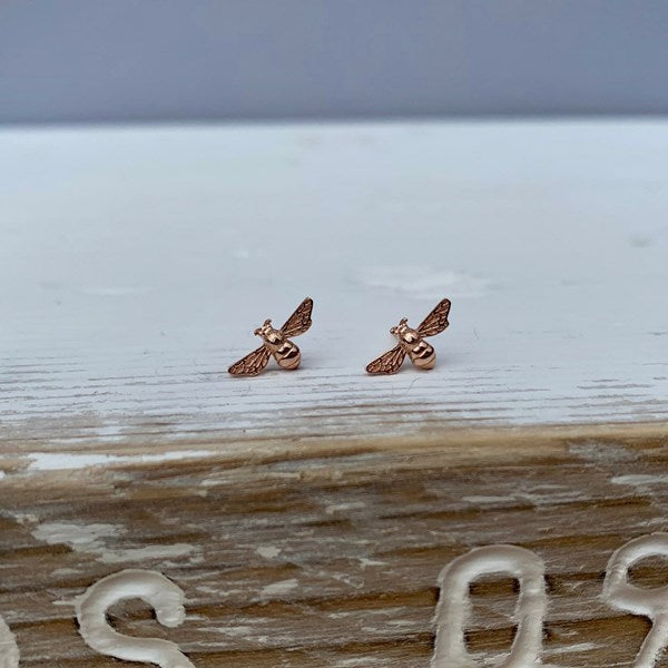A pair of small rose gold bee stud earrings resting on a rustic wooden surface with a light gray background.