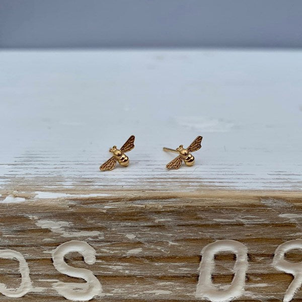 A pair of small gold bee stud earrings resting on a weathered wooden surface with a light gray background.