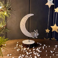 A decorative tabletop decoration featuring a crescent moon and sleeping rabbit silhouette in a speckled light grey finish mounted on a small round base, styled on a wooden surface with scattered white snow and holiday decor in the background.