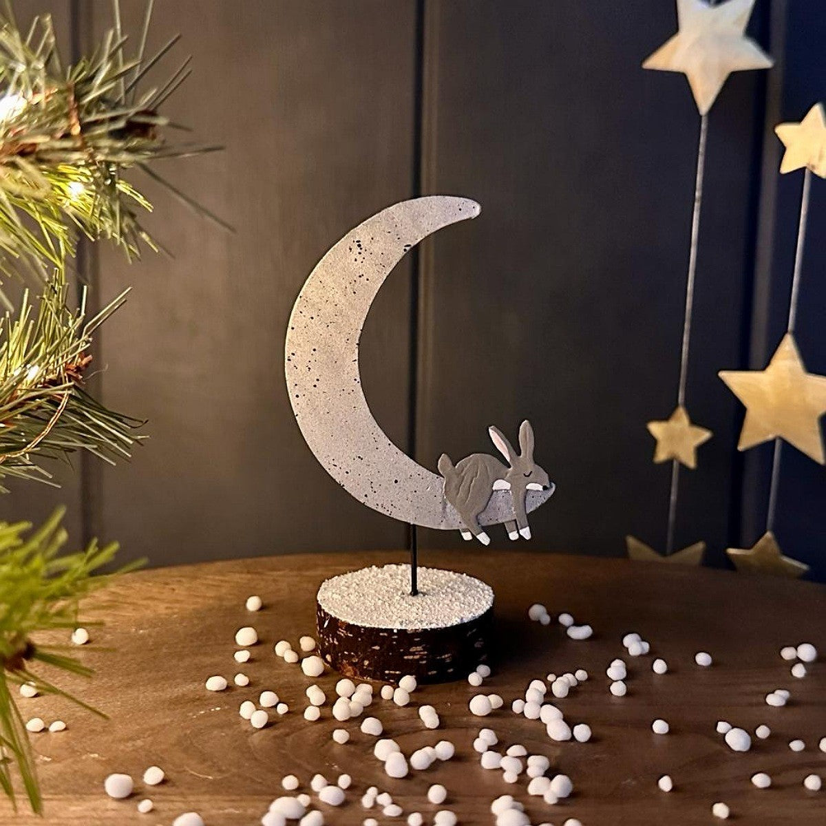 A decorative tabletop decoration featuring a crescent moon and sleeping rabbit silhouette in a speckled light grey finish mounted on a small round base, styled on a wooden surface with scattered white snow and holiday decor in the background.