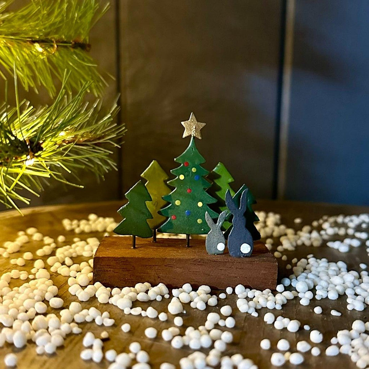 Handcrafted wooden Christmas tabletop decoration featuring three green trees and two grey rabbits on a small wooden base, styled on a wooden surface with white decorative beads and a blurred pine branch with warm lights.