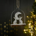 A white Christmas metal decoration featuring a crescent moon and two rabbits on a rustic wooden base, hanging by a twine loop against a dark panelled wall with a softly blurred lit Christmas tree in the background.