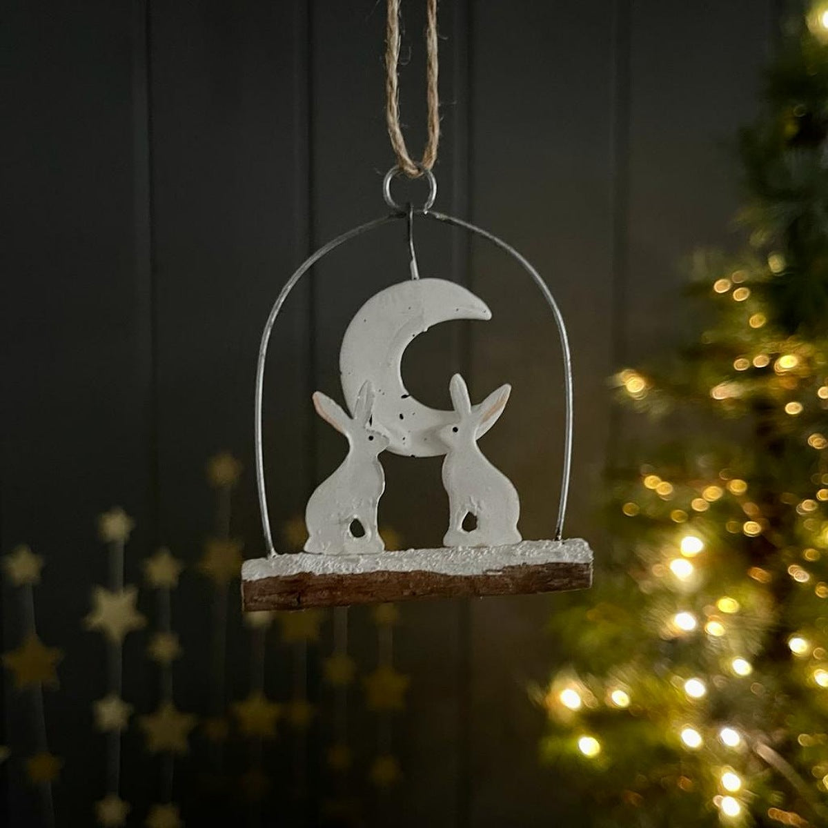 A white Christmas metal decoration featuring a crescent moon and two rabbits on a rustic wooden base, hanging by a twine loop against a dark panelled wall with a softly blurred lit Christmas tree in the background.