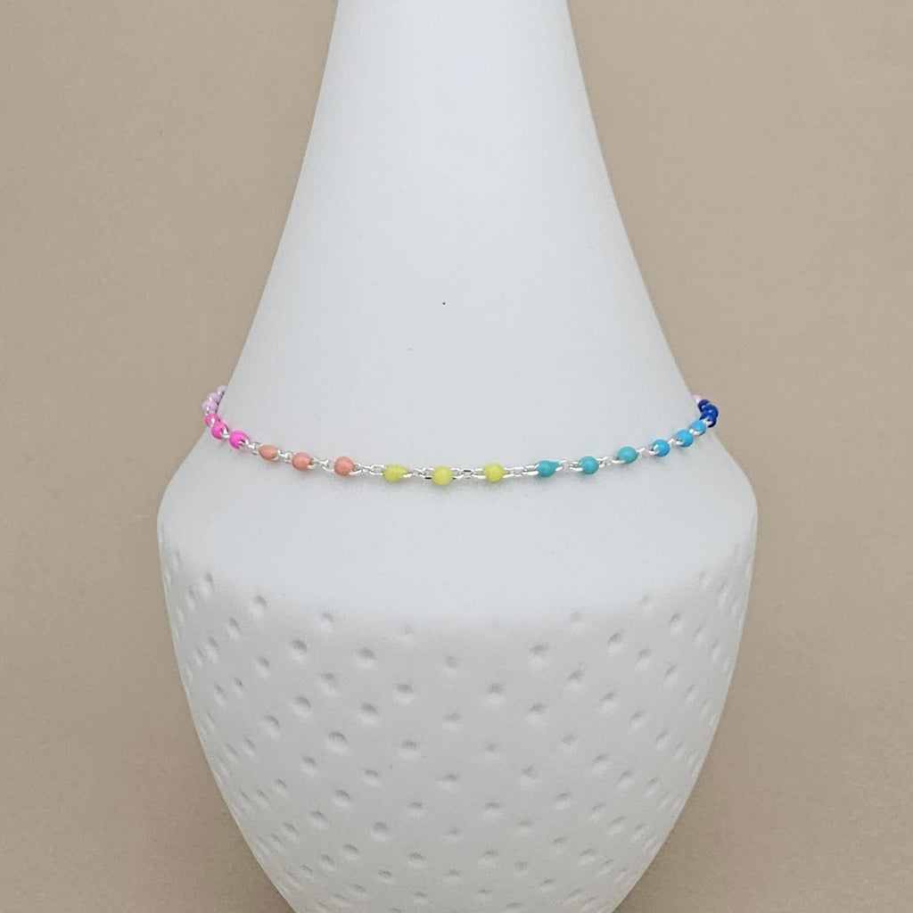 A delicate beaded anklet with a rainbow of small beads in pink, orange, yellow, green, blue, and purple, displayed on a white textured cone stand against a solid beige background.