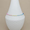 A delicate beaded anklet with a rainbow of small beads in pink, orange, yellow, green, blue, and purple, displayed on a white textured cone stand against a solid beige background.