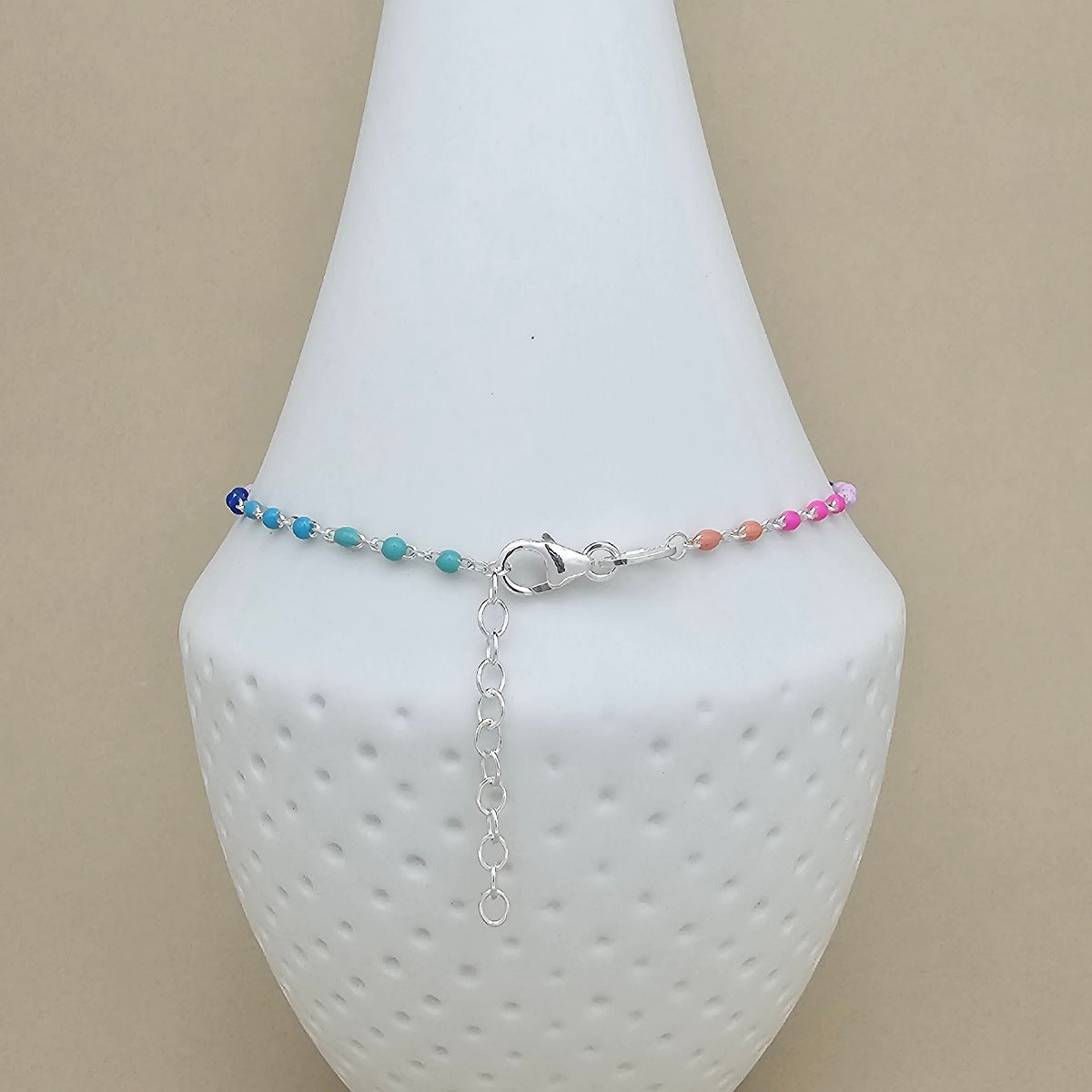 A delicate beaded anklet with blue, turquoise, and pink beads on a silver chain, displayed on a white textured jewelry bust against a beige background.
