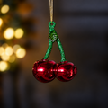 A cherry-shaped glass Christmas ornament with two shiny red bell-shaped cherries and glittery green stems, hanging from a gold string against a dark blurred background with warm bokeh lights.
