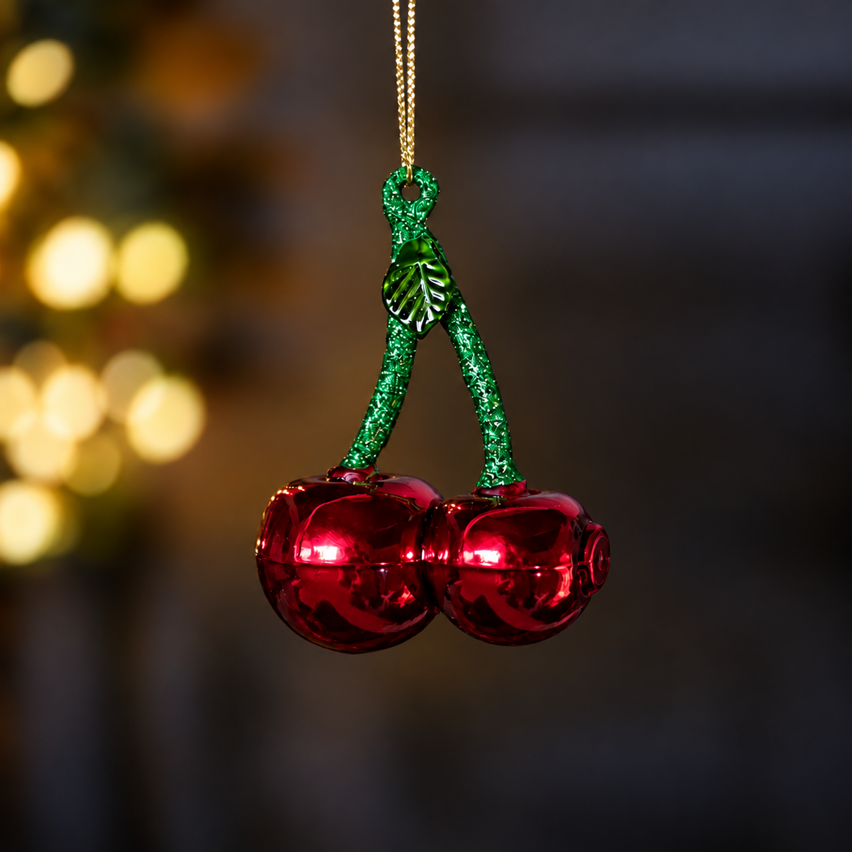 A cherry-shaped glass Christmas ornament with two shiny red bell-shaped cherries and glittery green stems, hanging from a gold string against a dark blurred background with warm bokeh lights.