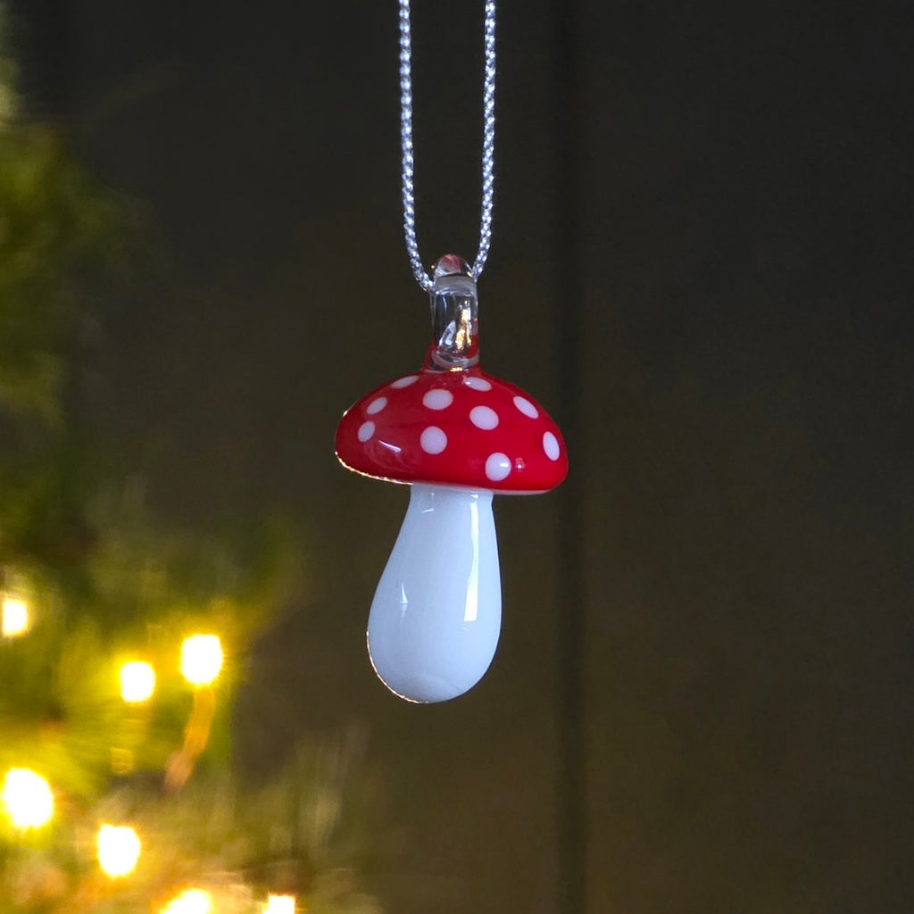 A mushroom-shaped glass hanging decoration with a glossy red cap, white polka dots, and a white teardrop-shaped body, hanging from a silver thread against a dark, softly blurred background with warm lights.