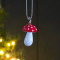 A mushroom-shaped glass hanging decoration with a glossy red cap, white polka dots, and a white teardrop-shaped body, hanging from a silver thread against a dark, softly blurred background with warm lights.