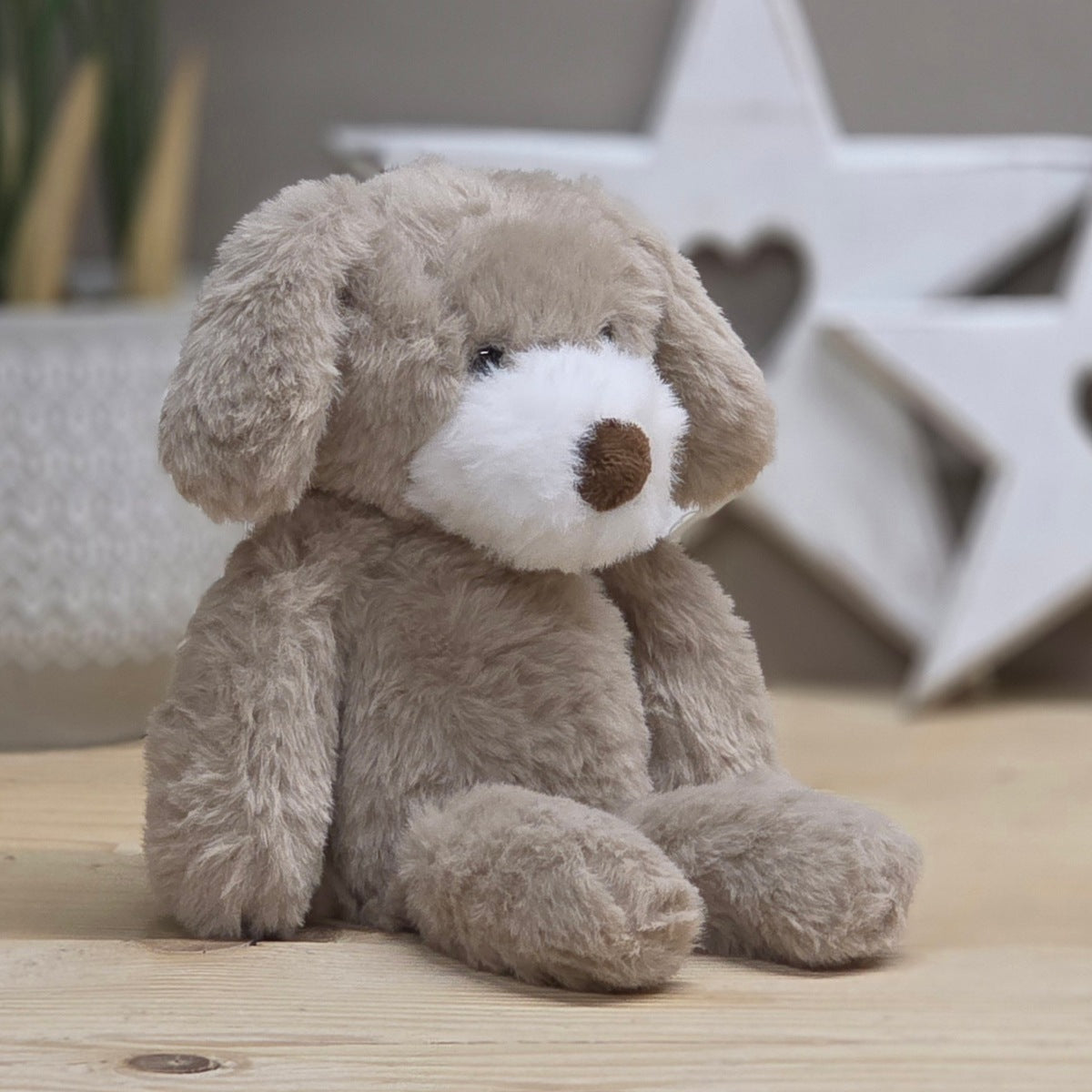 A beige soft fuzzy plush dog toy, with black eyes, white snout and brown nose, on a wooden surface with a blurred star-shaped object in the background
