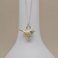 A two-tone bird pendant necklace on a delicate silver chain, displayed on a white jewelry bust against a soft beige background.