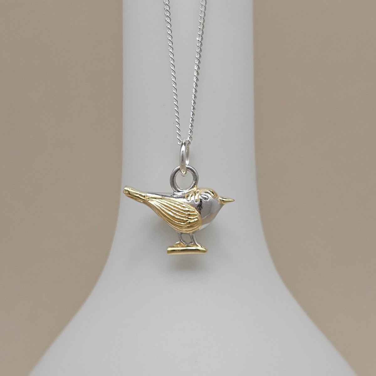 A two-tone bird pendant necklace on a delicate silver chain, displayed on a white jewelry bust against a soft beige background.