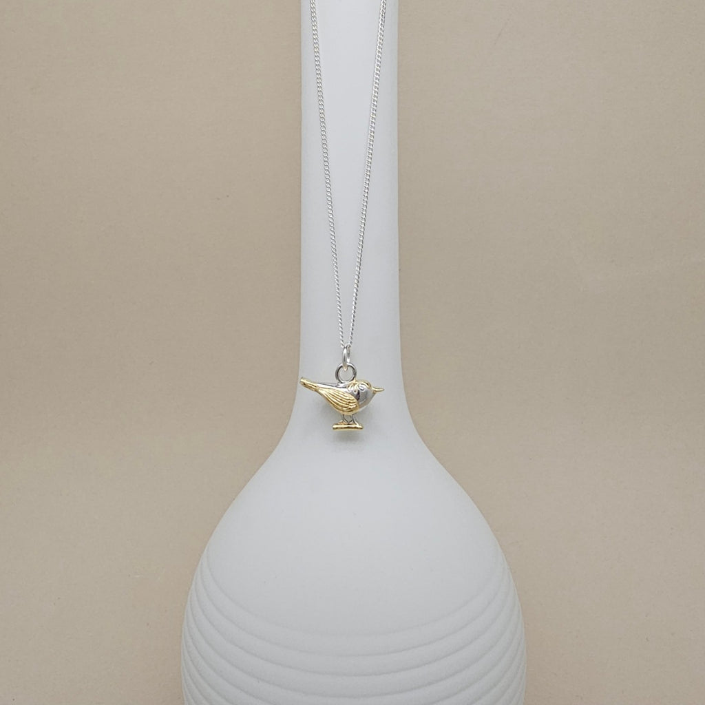 A silver chain necklace with a small bird pendant in gold-tone metal, displayed on a white ribbed vase against a beige background.