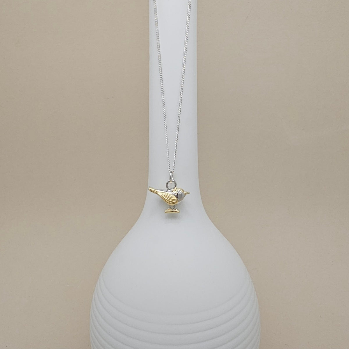 A silver chain necklace with a small bird pendant in gold-tone metal, displayed on a white ribbed vase against a beige background.