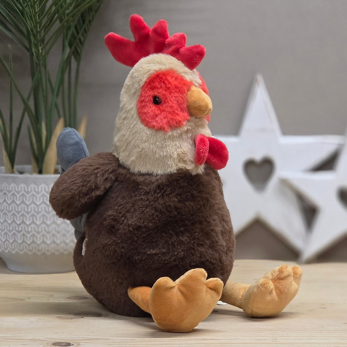 A plump brown, beige, red soft fuzzy plush rooster toy with an orange beak and legs, with black eyes.  On a wooden surface with a blurred star-shaped decorative objects in the background

