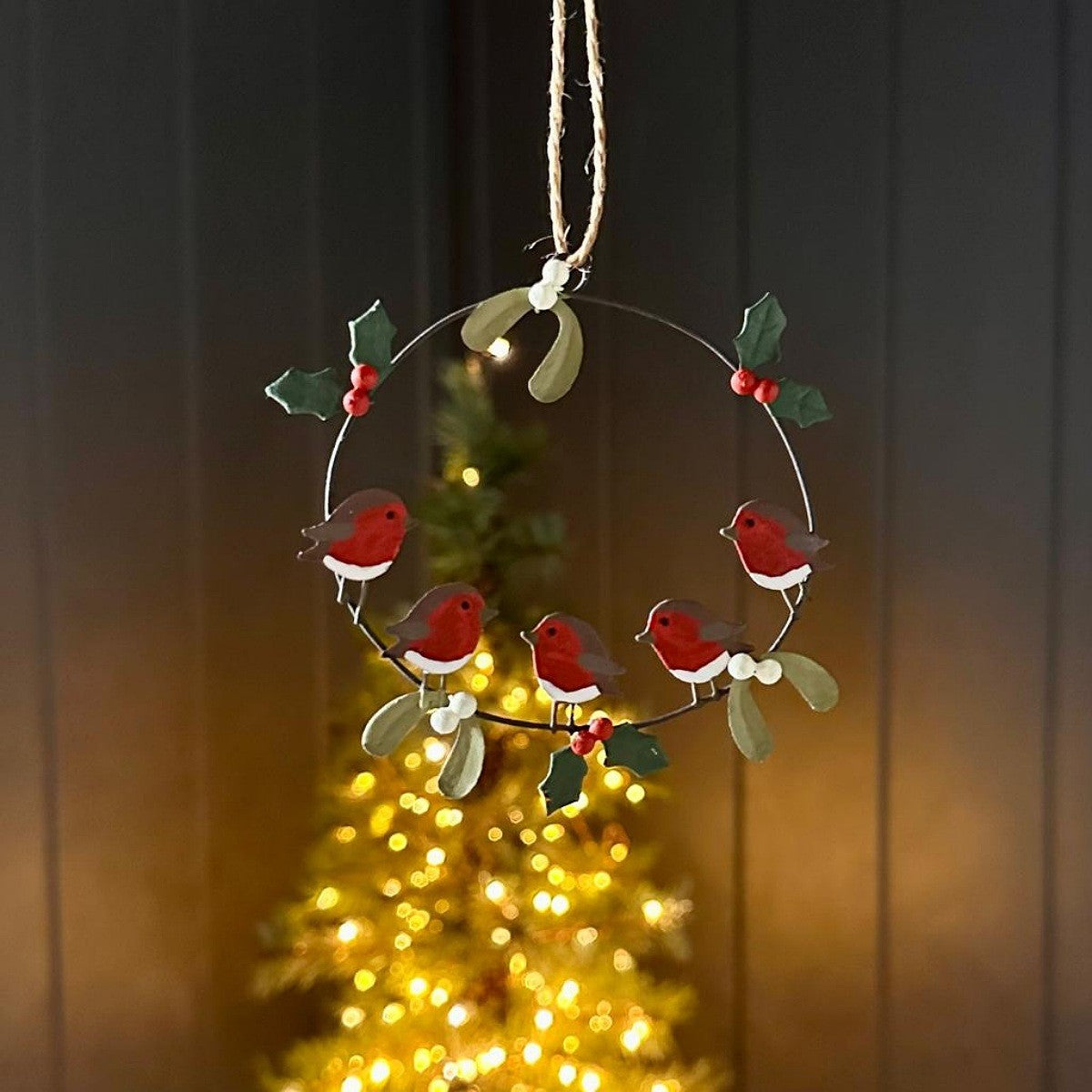 A hanging Christmas wreath decoration made of a thin metal ring with four red robin birds, green holly leaves, and small red berries. The wreath hangs from a rope against a dark panelled background with a blurred golden Christmas tree lights scene behind it.