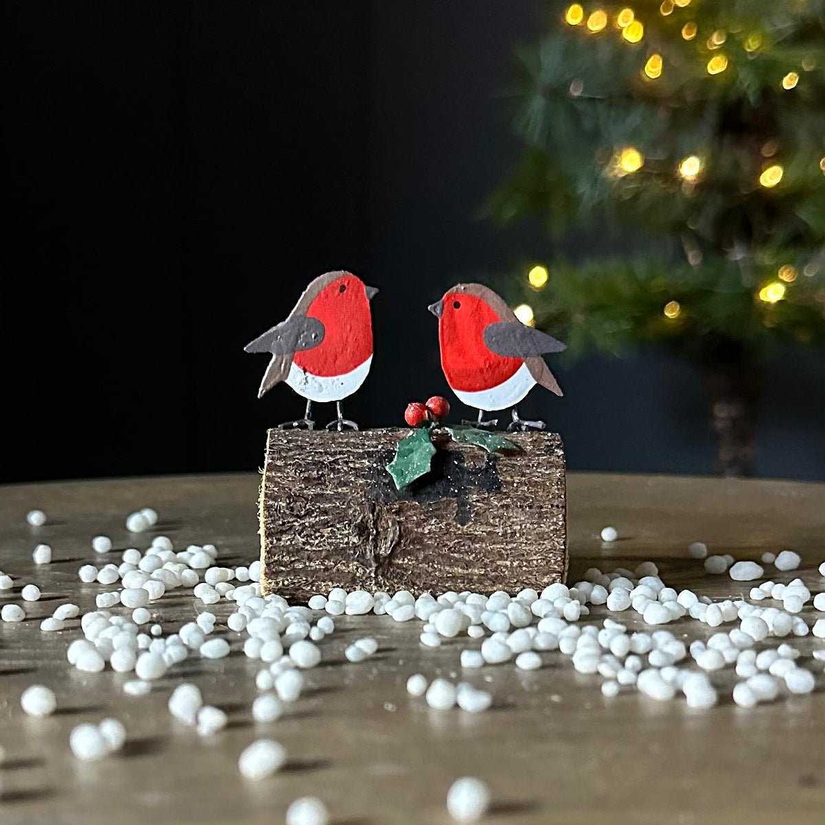 A decorative holiday tabletop scene with two small robin birds perched on a log-shaped base, surrounded by white stones and a blurred Christmas tree with warm lights in the background.
