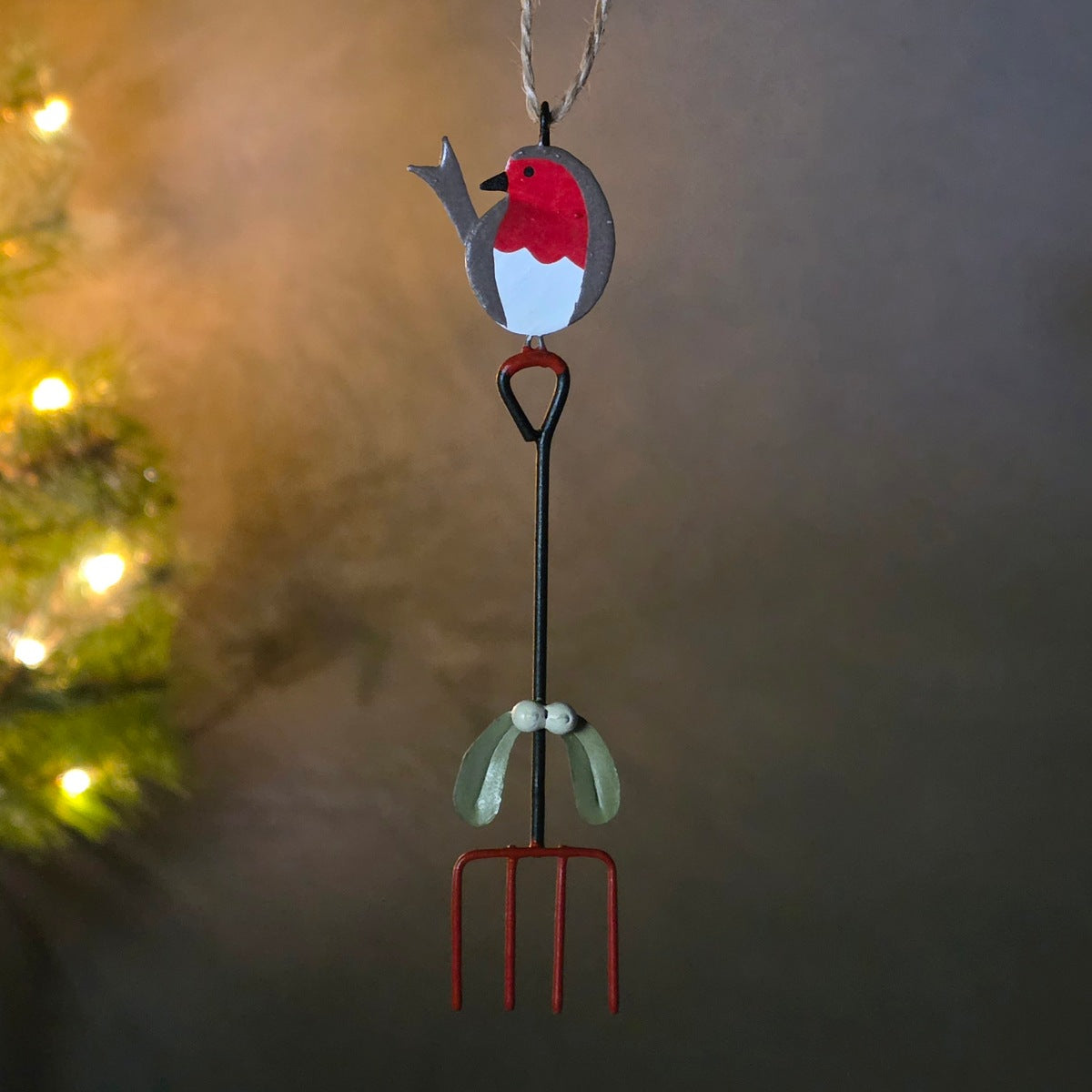 A Christmas robin bird ornament hanging from a metal garden fork. The robin has a red breast, gray back, and white belly, with a small hanging loop at the top.