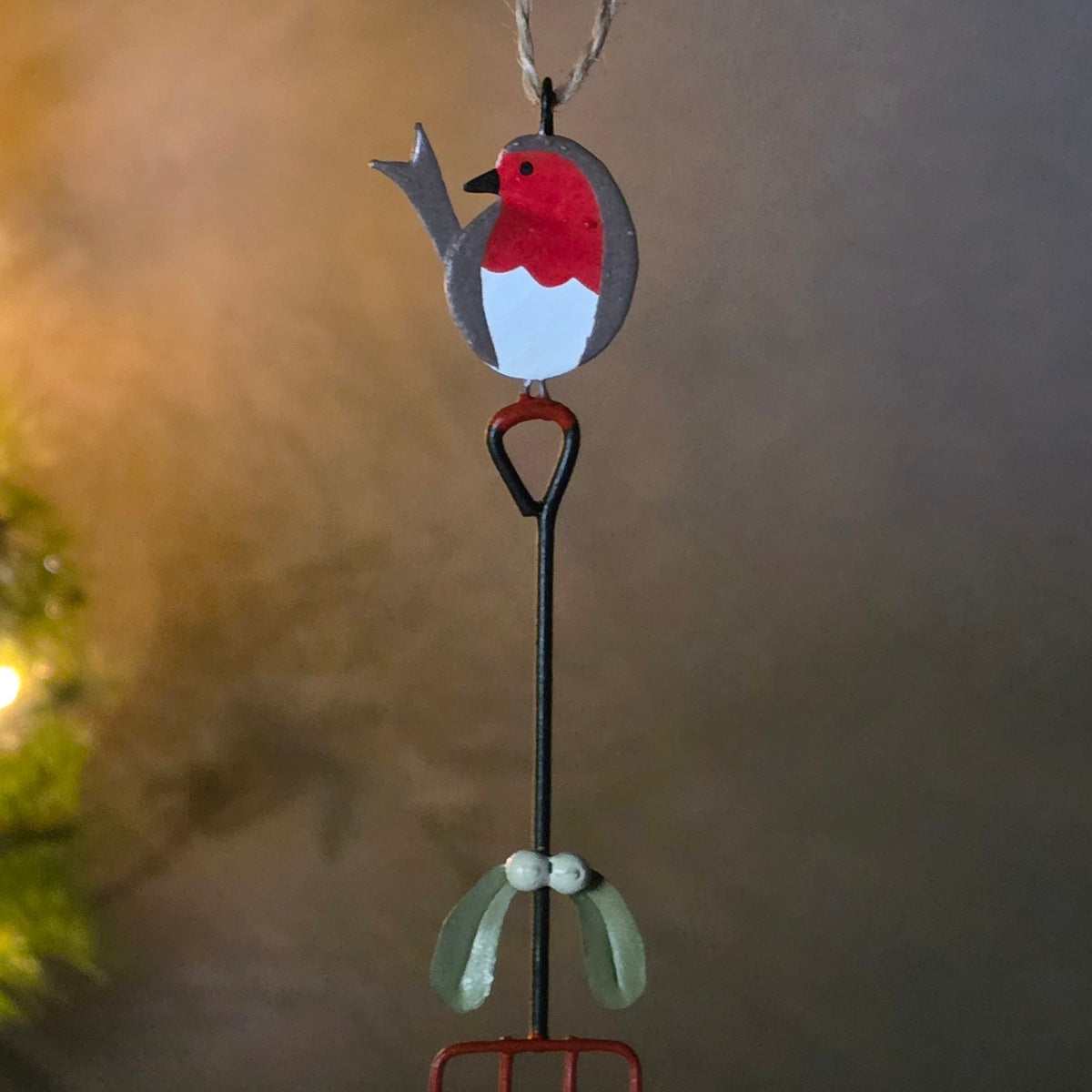 A decorative hanging ornament shaped like a robin bird perched on a miniature garden rake. The robin has a red chest, white belly, and gray back, with a small hanging loop at the top.