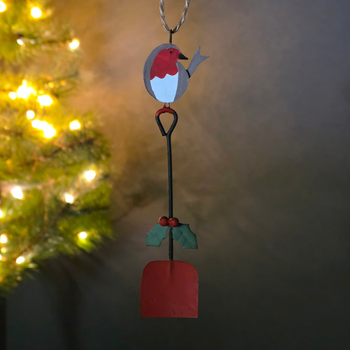 A hanging Christmas ornament shaped like a robin perched on a shovel, with a blurred Christmas tree and warm lights on the left side of the frame.